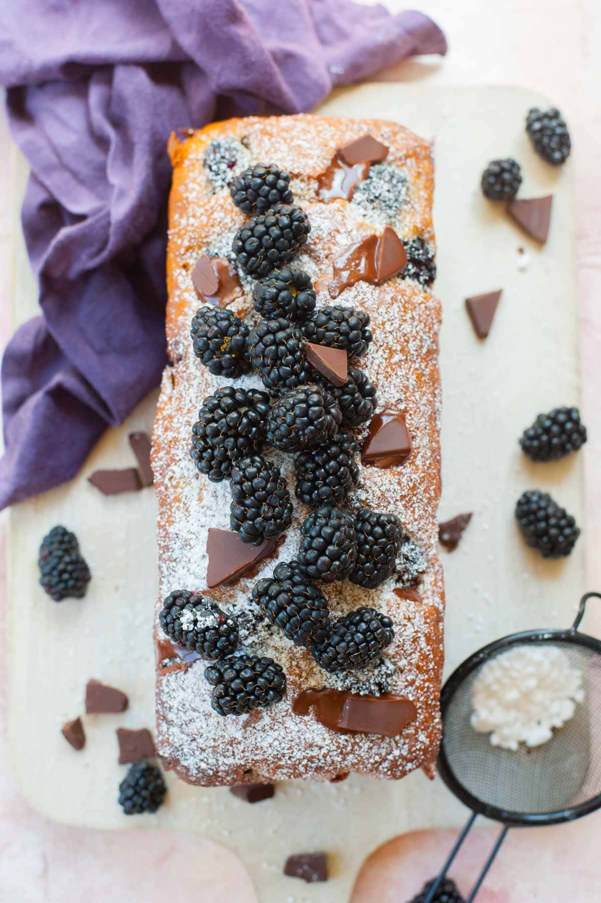 Blackberry banana bread with chocolate on a wooden board topped with fresh blackberries and chocolate pieces.