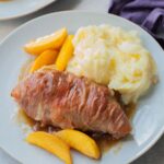 Prosciutto-wrapped chicken with peaches and mashed potatoes on a blue plate.