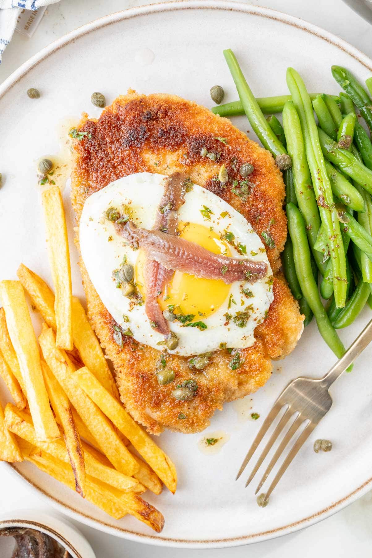 Schnitzel Holstein topped with a sunny-side-up egg, anchovy fillets, and capers on a plate with green beans and French fries.