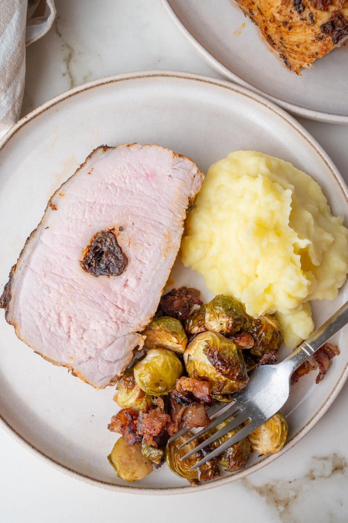 A slice of pork loin roast stuffed with prunes, mashed potatoes, and Brussels sprouts with bacon on a beige plate.