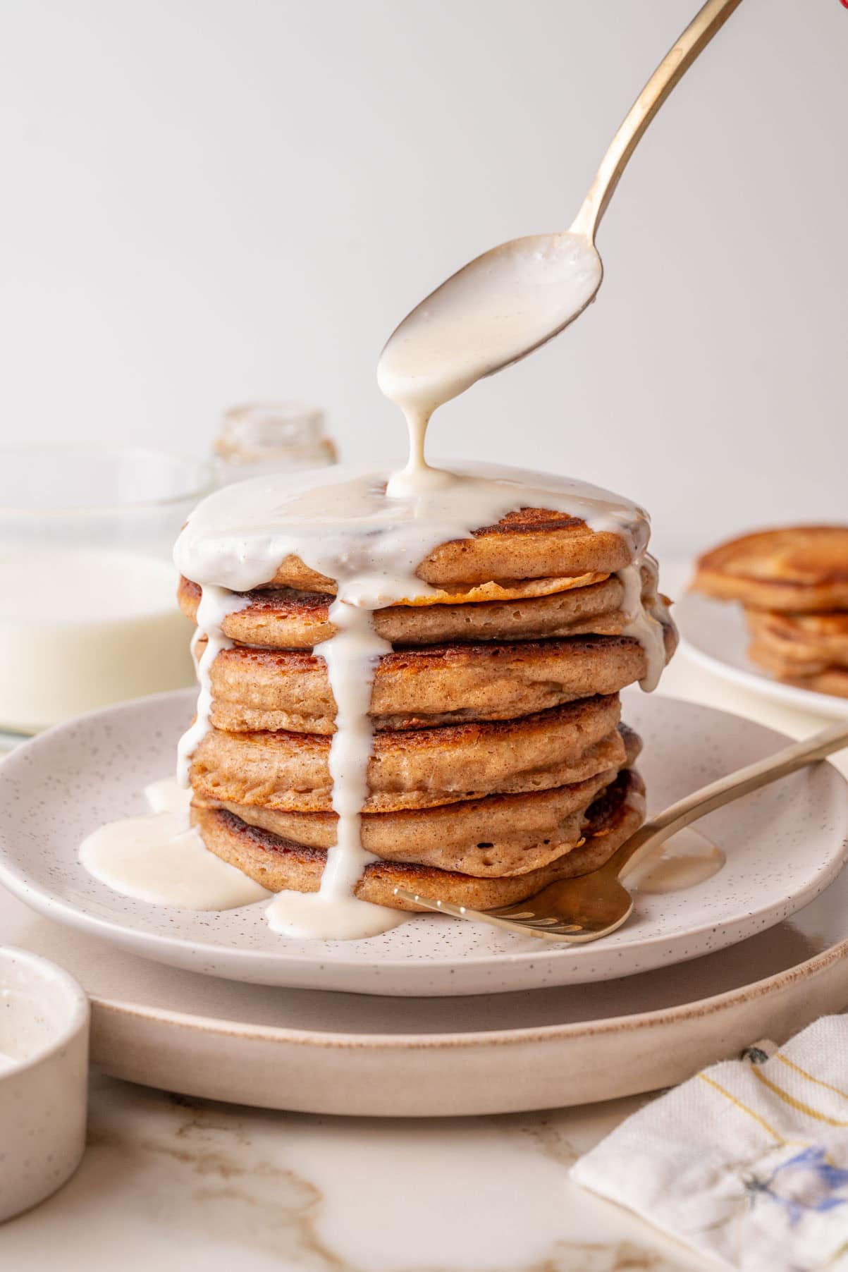 A stack of golden-brown cinnamon pancakes on a white speckled plate, with a spoon drizzling creamy glaze over the top. The glaze is dripping down the sides of the pancakes, and a gold fork rests beside the stack.