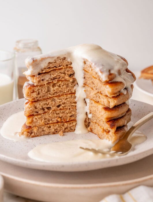 A tall stack of fluffy cinnamon pancakes with a generous cream cheese glaze dripping down the sides, served on a white speckled plate with a fork and extra glaze on the side. A large wedge is cut out, showing the soft, airy texture inside.