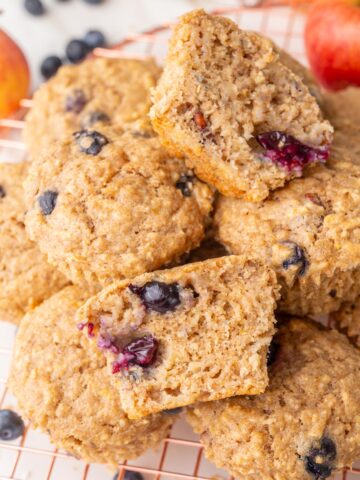 Freshly baked healthy breakfast muffins stacked on a wire cooling rack, with one muffin cut in half to show its soft and moist texture with visible blueberries inside.