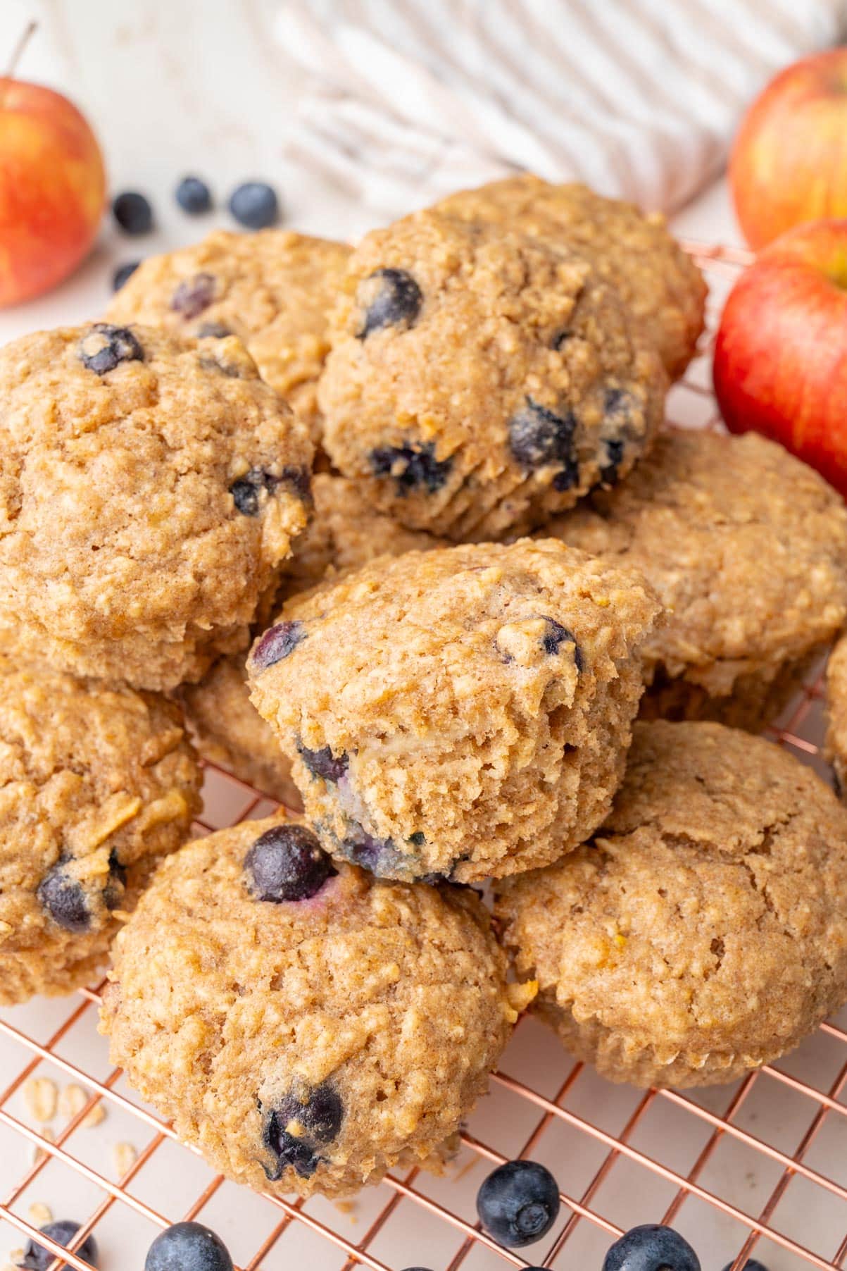 A pile of golden-brown healthy breakfast muffins on a wire cooling rack, surrounded by fresh blueberries and apples in the background.