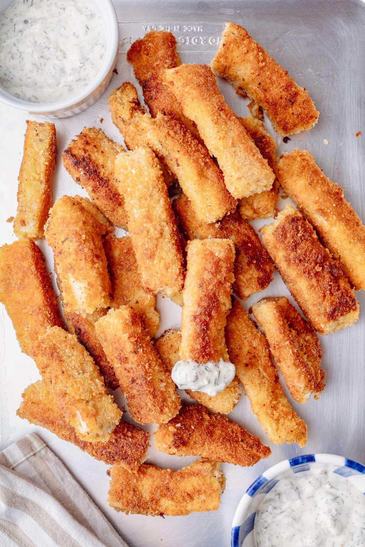 A baking sheet filled with golden-brown homemade fish sticks served with tartar sauce.