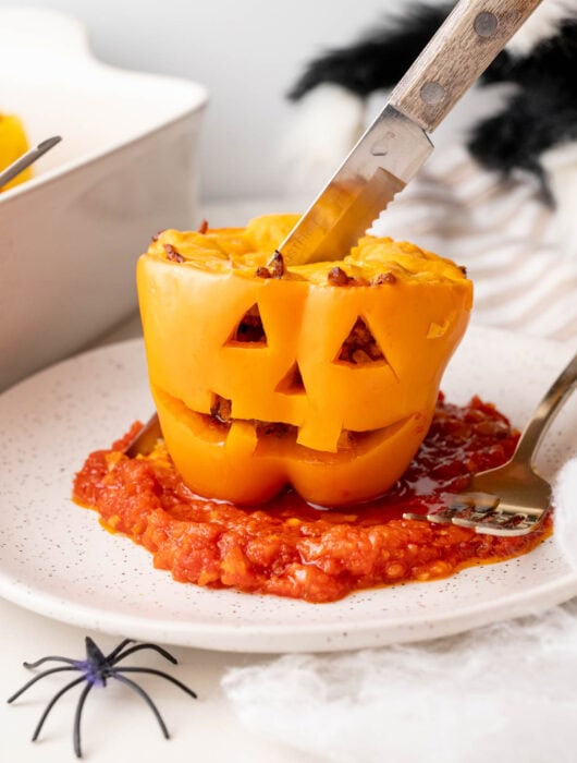 Stuffed pepper with carved face served on tomato sauce with knife cutting into the top.