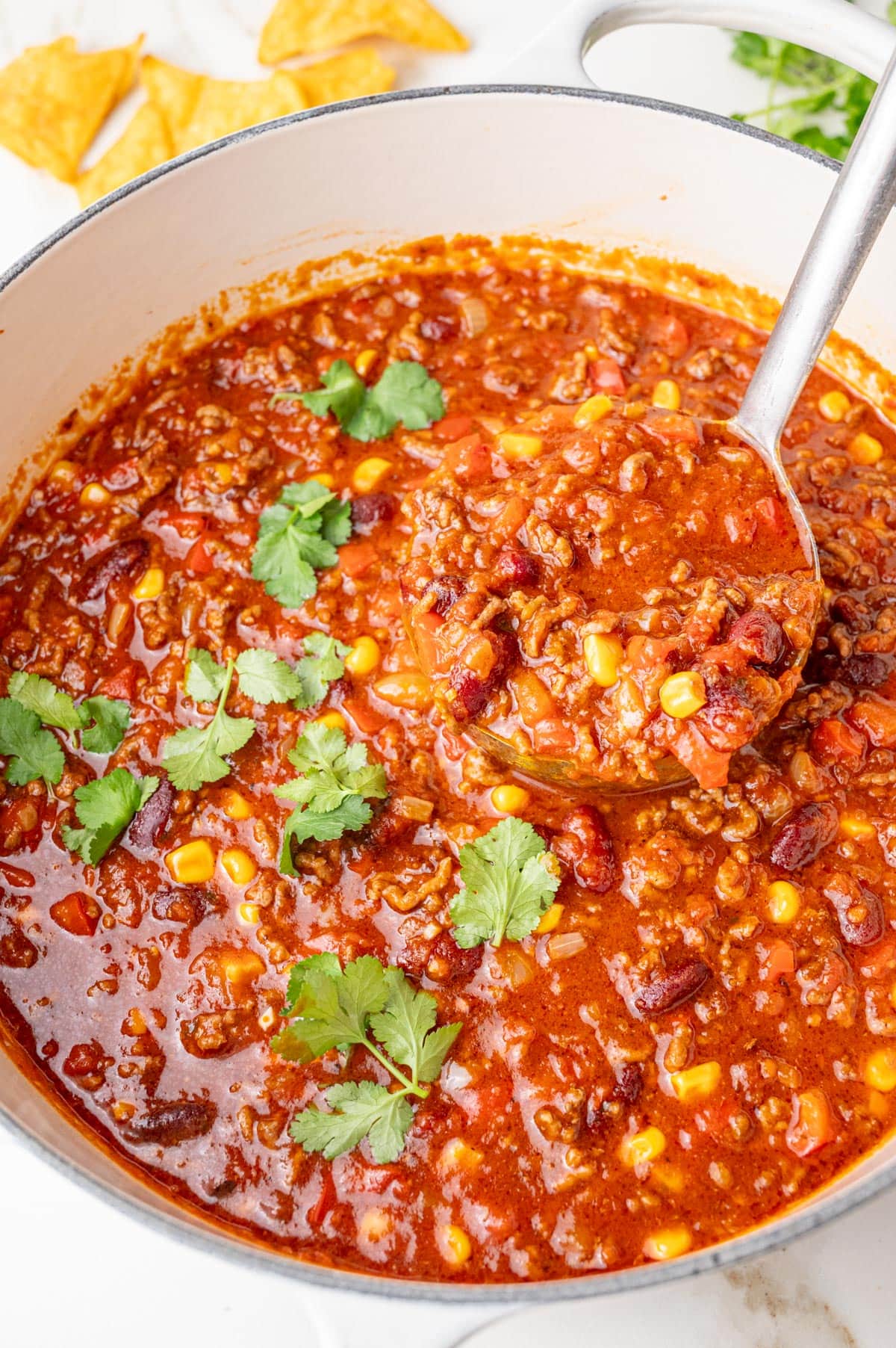 Hearty chili con carne with ground beef, beans, corn, and tomatoes simmering in a white Dutch oven, garnished with fresh cilantro and served with tortilla chips.