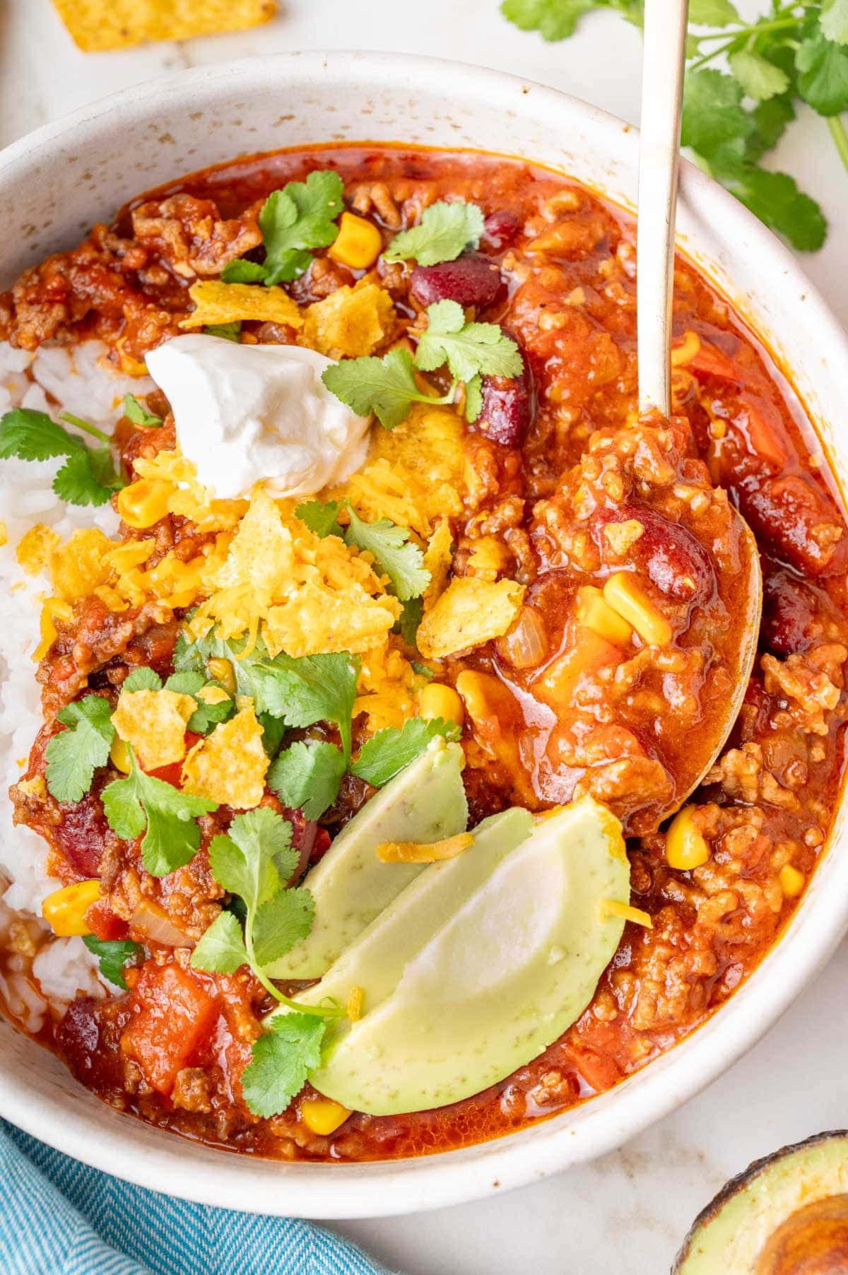 Bowl of chili con carne served over rice, topped with sour cream, avocado slices, cheddar cheese, crushed tortilla chips, and cilantro.