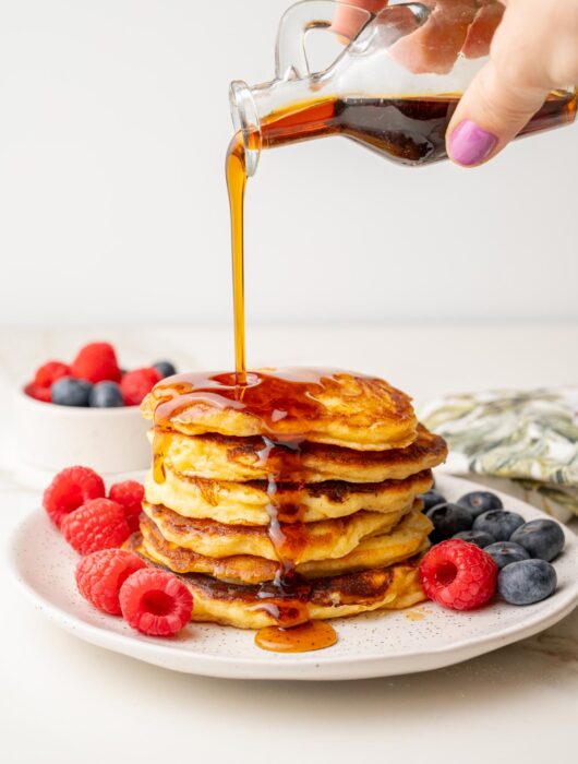 A stack of yogurt pancakes on a plate with barries. Maple syrup is being poured over.