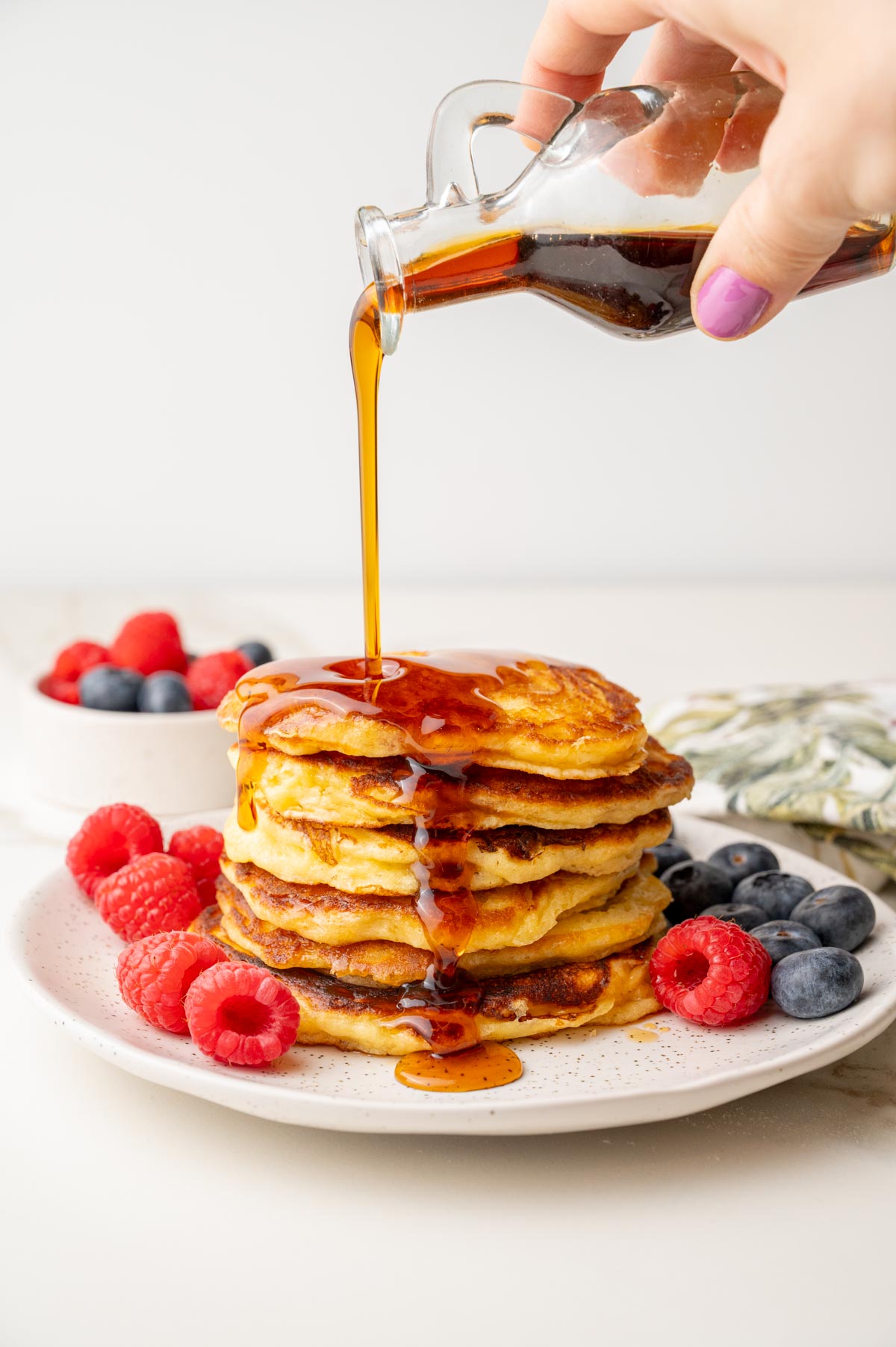Maple syrup being poured over a stack of fluffy yogurt pancakes, served with raspberries and blueberries.