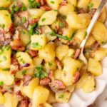 German potato salad topped with bacon and chives in a beige bowl.