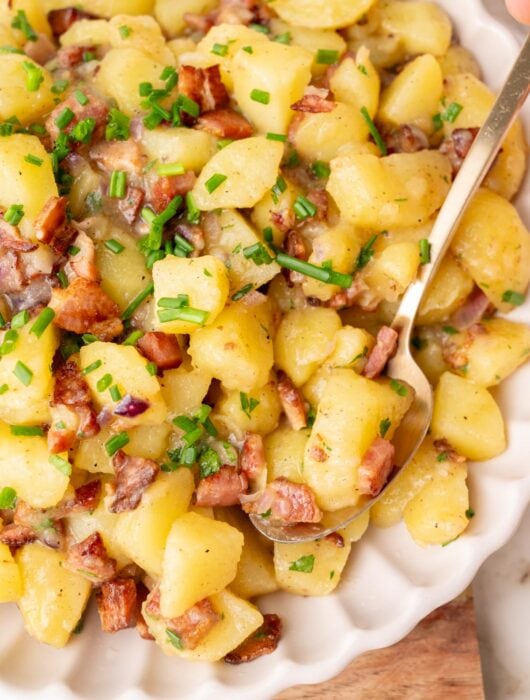 German potato salad topped with bacon and chives in a beige bowl.