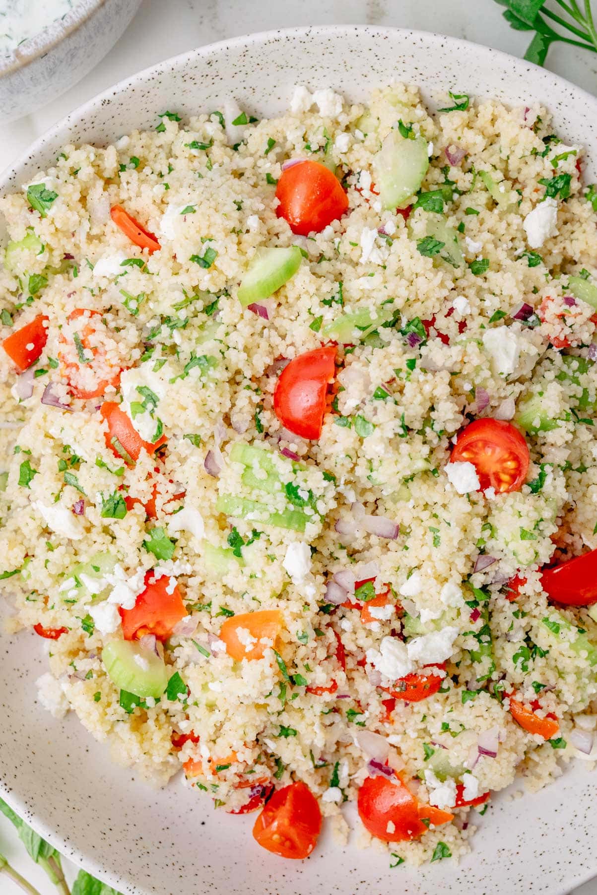 Couscous salad with cherry tomatoes, cucumber, red onion, feta, parsley, and mint in a white bowl.